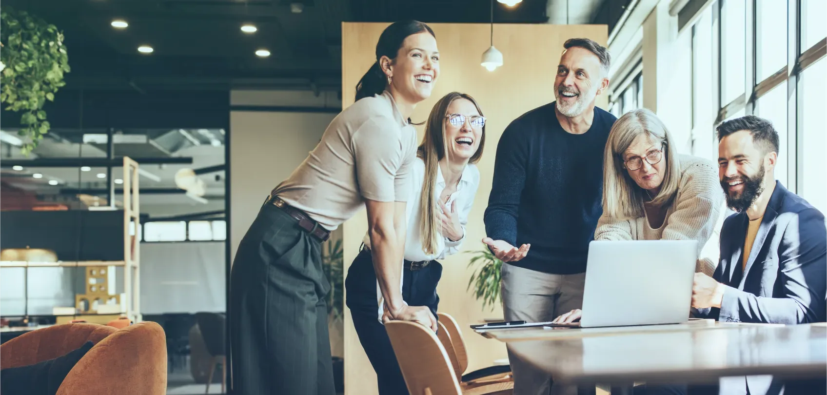 Diverse team gathered around a laptop, smiling and collaborating in a modern office space.