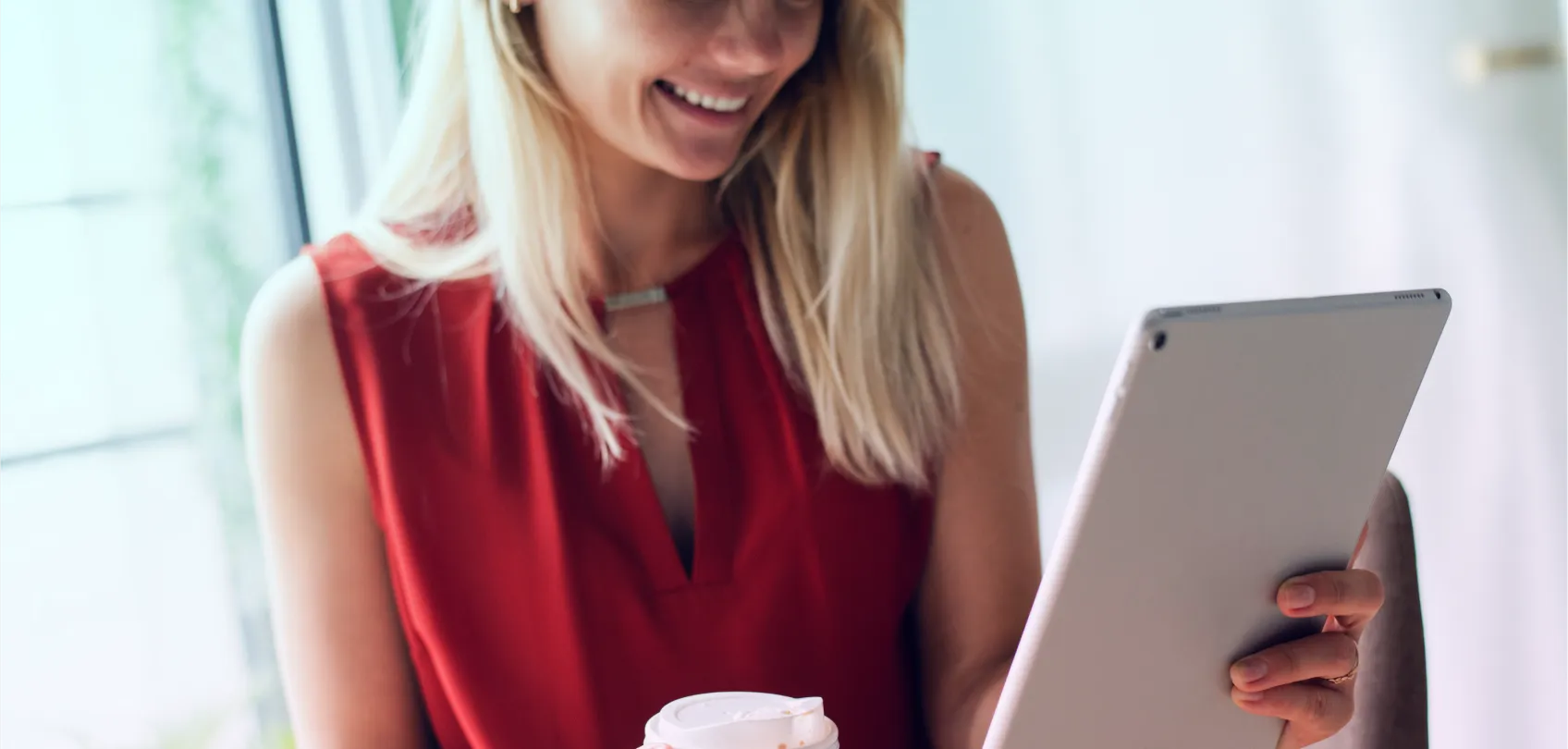 Woman smiling while reading on a tablet and holding a cup of coffee, in a bright setting.
