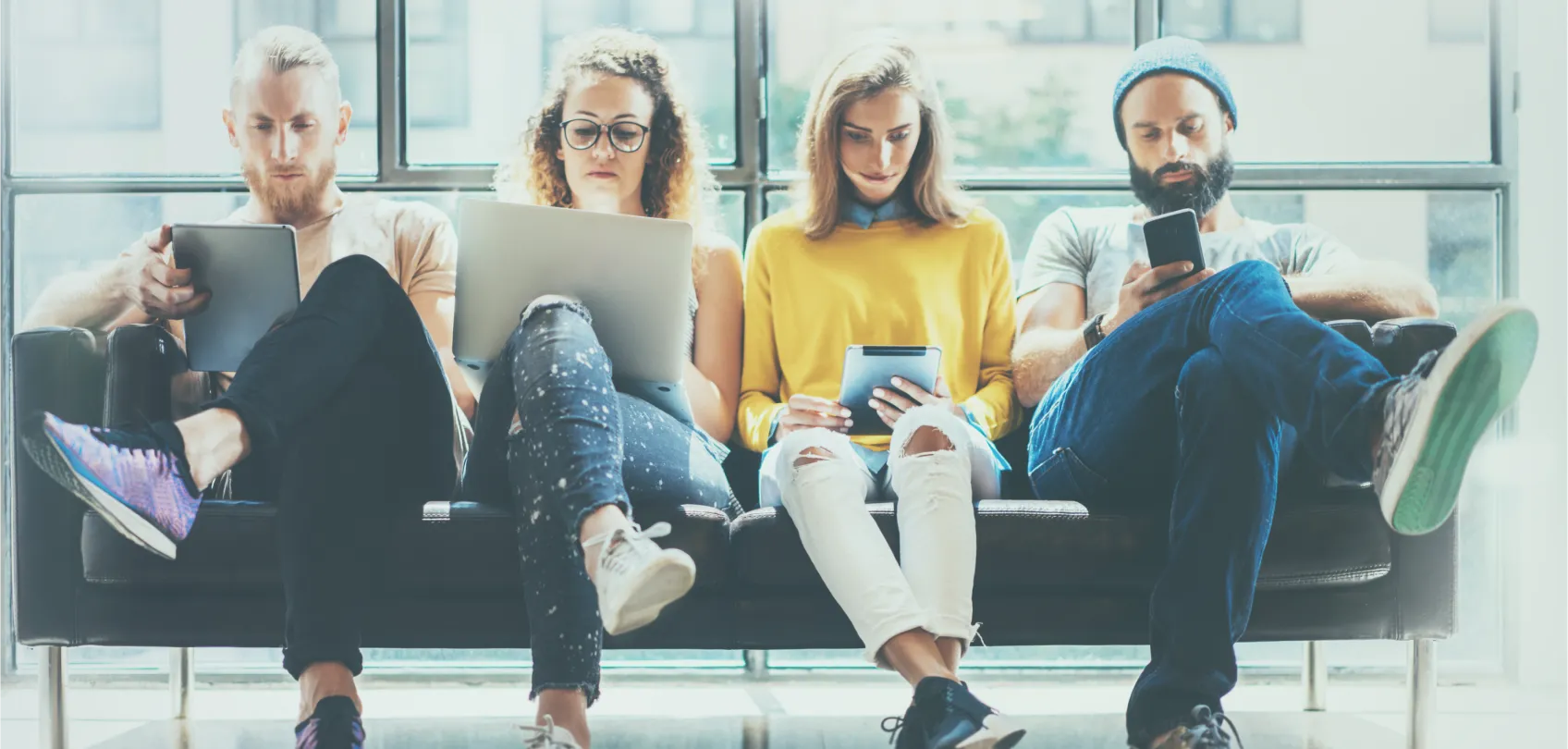 Four young people sitting on a sofa, each using a device in a bright space, representing the use of technology and data in marketing.