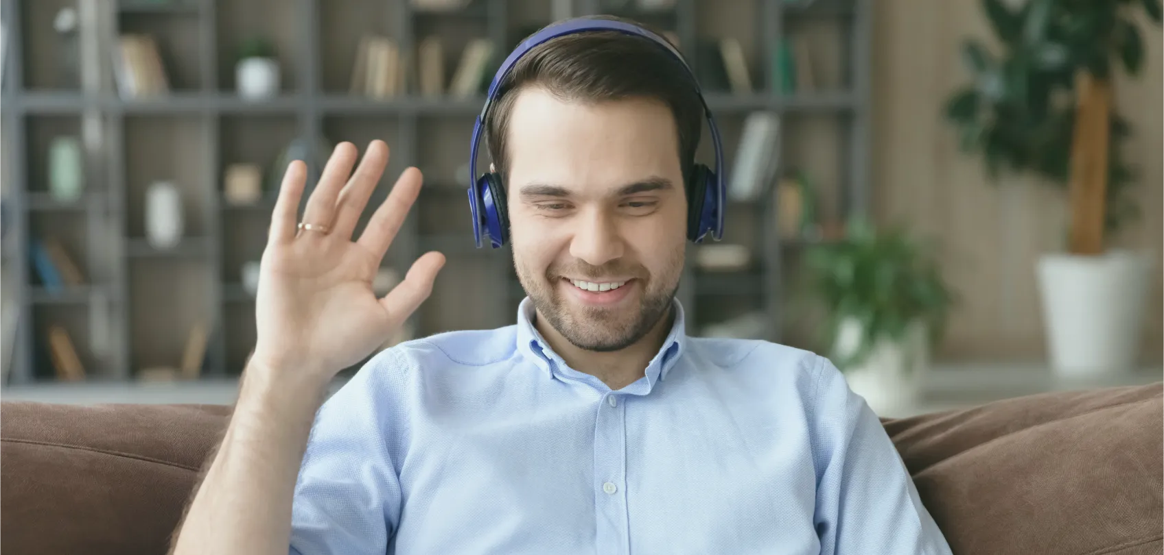 Young man with blue headphones waving on a video call from home, with a bookshelf and plants in the background.
